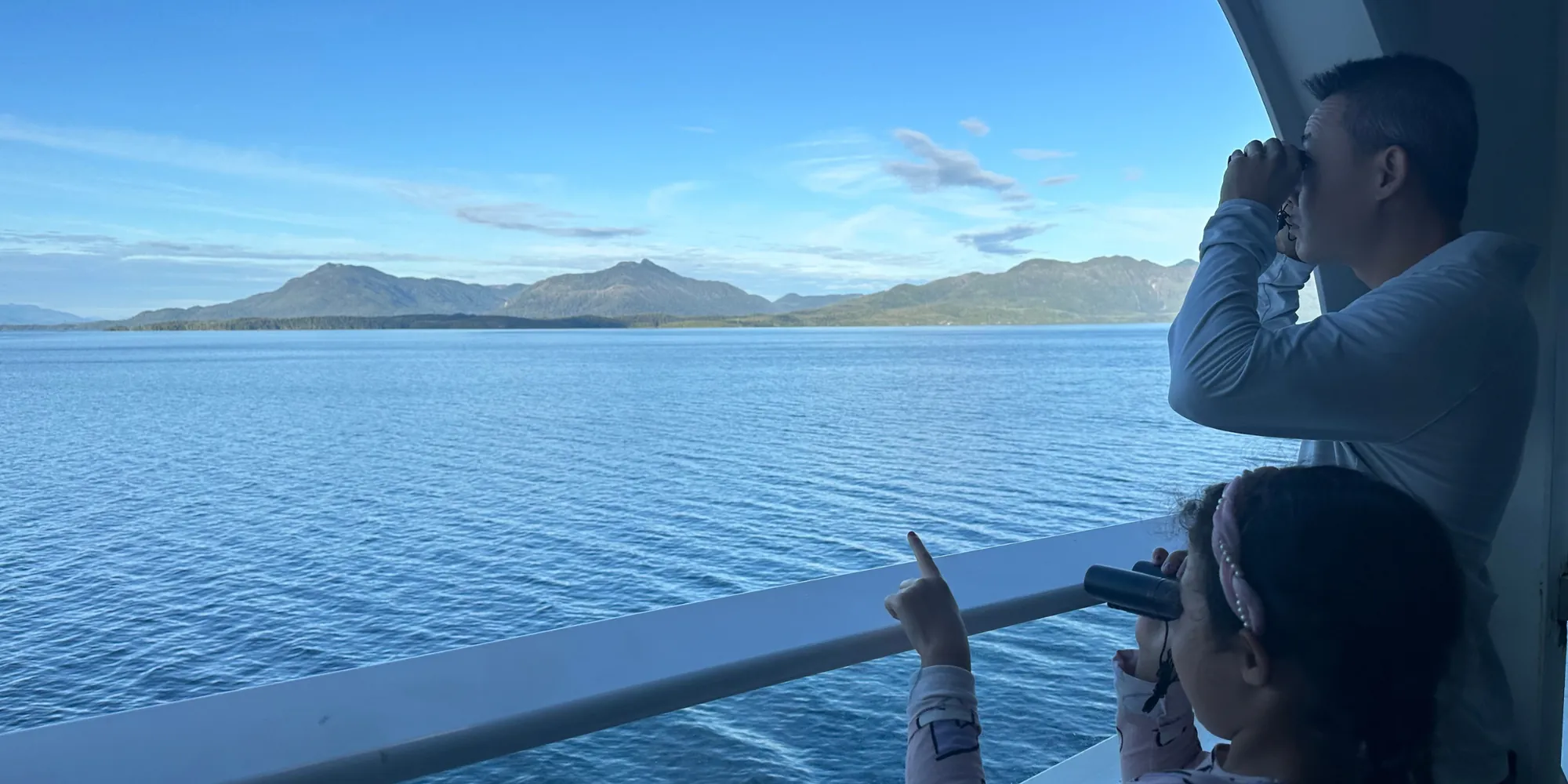 A girl and her dad looking through binoculars from a cruise ship balcony at green mountains across calm water