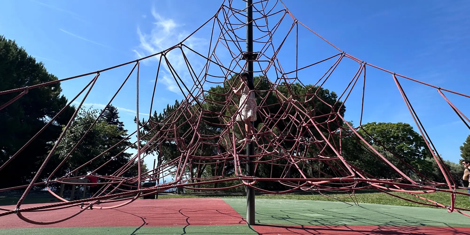 A girl climbing a giant red rope net play structure in a sunny park near the port